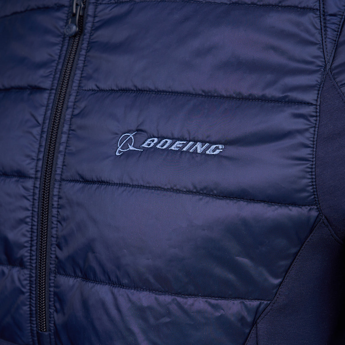 Close-up of a navy blue puffer jacket with the Boeing logo embroidered in light blue on the chest. The jacket's texture is glossy and quilted.